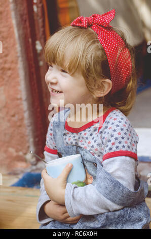 White cute toddler girl wearing bright red bandana on a had and retro style denim drinks cocoa sitting on a house threshold. Happy child portrait. Stock Photo