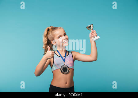 Cheerful little sports girl celebrating the win isolated over blue background, wearing a gold medal, showing a trophy Stock Photo