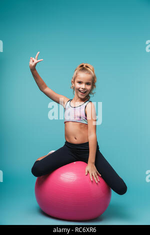 Cheerful little girl wearing sport clothes doing exercises with fitness ball isolated over blue background Stock Photo