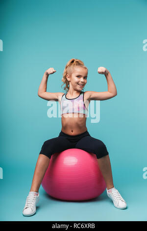 Cheerful little girl wearing sport clothes doing exercises with fitness ball isolated over blue background Stock Photo