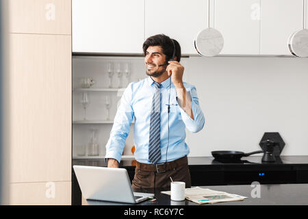 Confident young businessman working at home at the kitchen, using laptop computer, communicating through headphone set Stock Photo