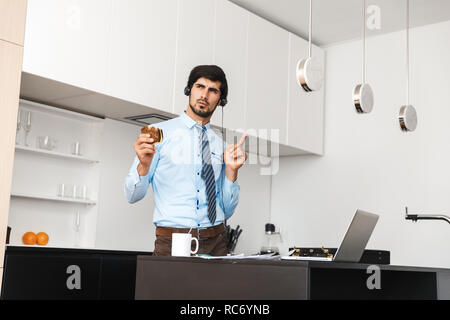 Image of a serious concentrated young business man at the kitchen eat sandwich using laptop computer wearing headphones. Stock Photo
