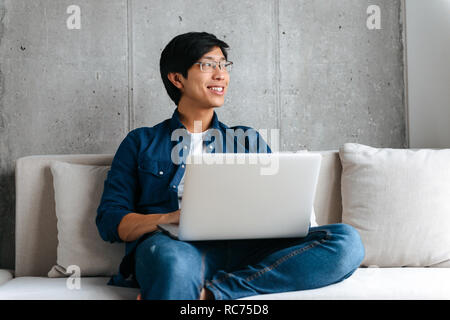 Smiling young asian man sitting on a couch with laptop computer at home Stock Photo