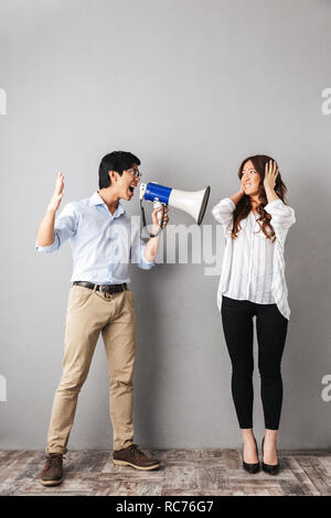 Angry asian business couple standing isolated over gray background, man holding loudspeaker and screaming Stock Photo