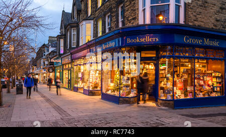 Golden glow of lights & attractive window display, exterior of The Grove Bookshop is welcoming as people walk in or past - Ilkley, West Yorkshire, UK. Stock Photo