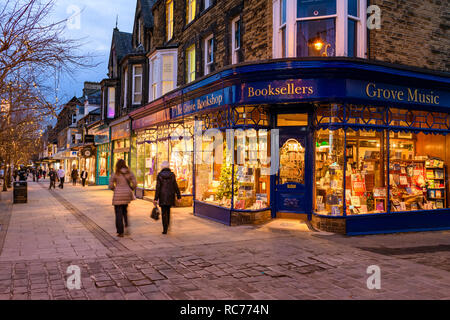 Golden glow of evening lights & attractive window display, exterior of The Grove Bookshop is welcoming (people walk past) - Ilkley, West Yorkshire, UK Stock Photo