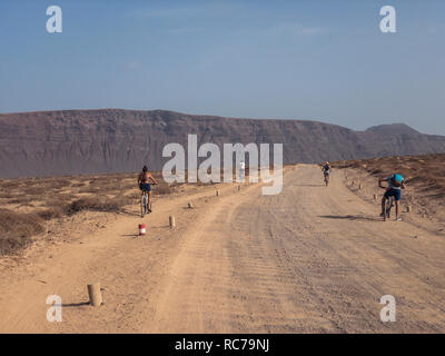 People cycling on a dirt path in the island of La Graciosa, Lanzarote, Canary Islands. Spain. In the background El risco, Lanzarote's mountain Stock Photo