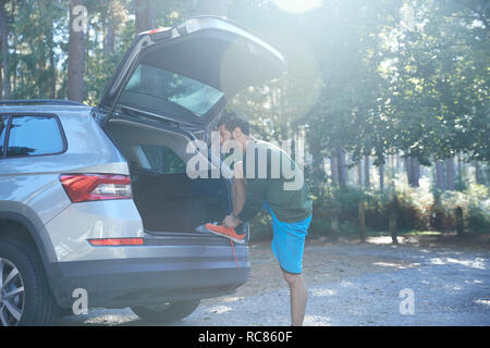 Male runner tying laces on car boot in sunlit forest Stock Photo