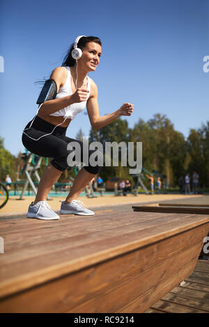 Cheerful woman in headphones doing jump squat on step Stock Photo