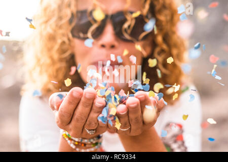 Close up of woman's habd and beautiful lady blowing out coloured carnival party confetti - focus on colors papers and hapiness and joyful lifestyle co Stock Photo