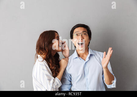 Excited asian couple standing isolated over gray background, telling secrets to each other Stock Photo