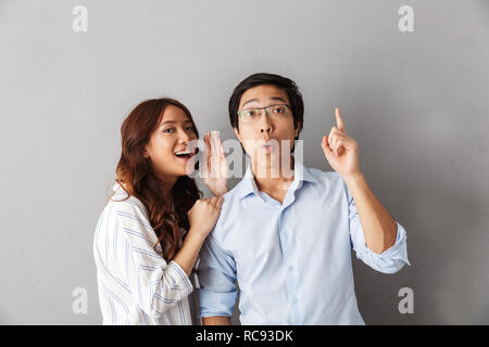 Excited asian couple standing isolated over gray background, telling secrets to each other Stock Photo