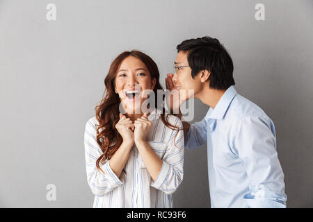 Excited asian couple standing isolated over gray background, telling secrets to each other Stock Photo