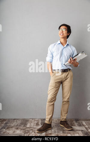 Full length of a smiling asian business man standing over gray background, holding folders with documents Stock Photo