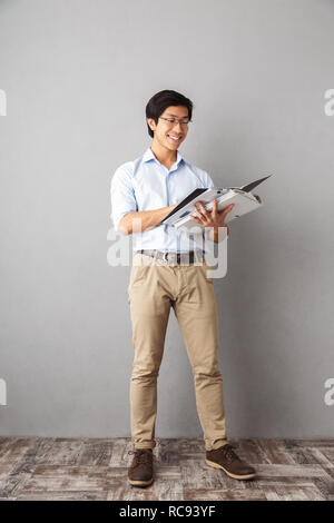 Full length of a smiling asian business man standing over gray background, holding folders with documents Stock Photo