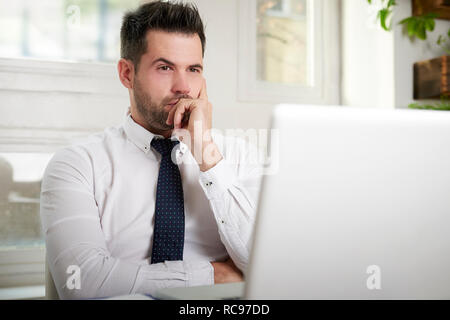 Portrait of careworn businessman sitting at office desk with hand on his chin while working on laptop. Stock Photo
