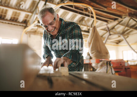 Happy senior male carpenter cutting wood planks on table saw machine. Smiling mature man working in carpentry workshop. Stock Photo