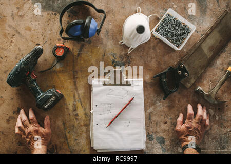 Top view of carpenter workbench with clipboard, drilling machine, mask, hand saw, hammer, screws, tape measure. POV of carpenter's workbench with tool Stock Photo