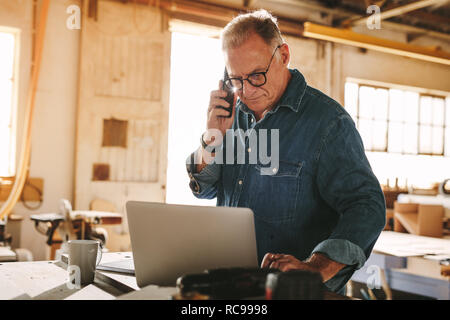 Senior man talking on cell phone and using laptop on work table. Mature carpenter working on laptop and answering phone call in his carpentry workshop Stock Photo