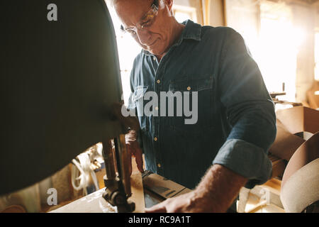 Senior male carpenter working on band saw machine cutting wood in his workshop. Carpenter cutting wood on machine at carpentry workshop. Stock Photo