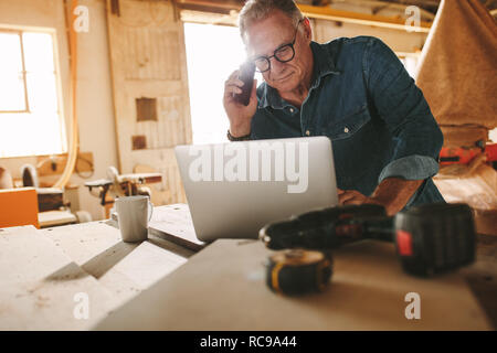 Senior man using laptop and speaking by smartphone in his carpentry workshop. Middle aged carpenter working on laptop and answering phone call in his  Stock Photo