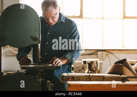Mature worker in the carpentry workshop cuts the wood using band saw.  Carpenter working in carpentry preparing furniture parts. Stock Photo