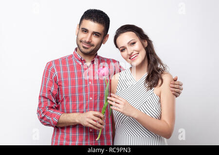 happy couple with tulip, smiling, hugging and looking at camera. Portrait of handsome man in red checkered shirt and beautiful woman in white striped  Stock Photo