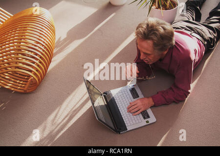 Mann mittleren Alters mit Notebook auf dem Boden liegend | middleaged man is lying on the floor with a notebook Stock Photo
