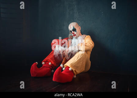 Tired bloody clown with baseball bat sitting on the floor. Man with makeup in carnival costume, crazy maniac Stock Photo