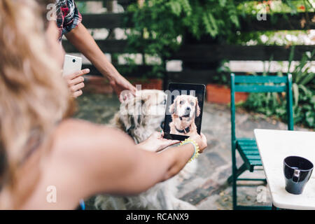 Rear view of a young smiling couple sitting on the steps outside the wooden hut taking a picture of their dog Stock Photo