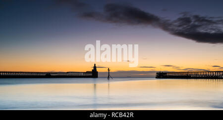 Winter sunrise at Blyth harbour looking across the mirror reflection on the tide towards the two silhouette piers and the lighthouse of the horizon Stock Photo