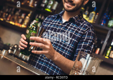 Young barman standing at bar counter holding jigger with alcohol close-up smiling cheerful blurred background Stock Photo