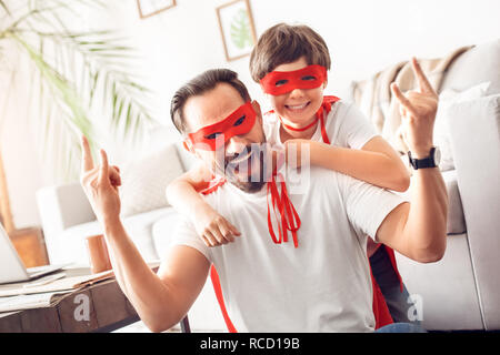 Father and little son wearing superheroe costumes together at home posing to camera man showing horns laughing cheerful Stock Photo