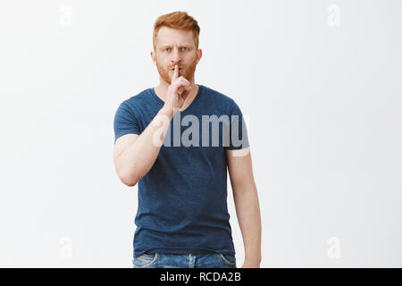 Shh, I need silence to focus on work. Portrait of handsome serious-looking and strict father with red hair and bristle, frowning, making shush gesture with index finger over mouth, asking keep quiet Stock Photo