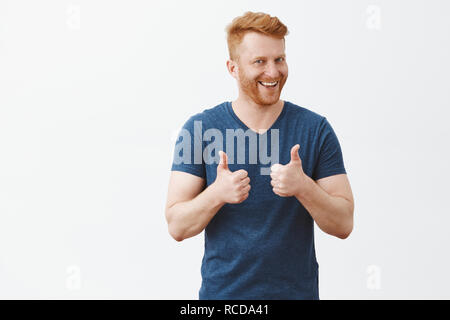 Nice job man, like it. Pleased good-looking happy male friend with ginger hair and brislte, showing thumbs up and smiling broadly, giving positive feedback, sharing his positive opinion over grey wall Stock Photo