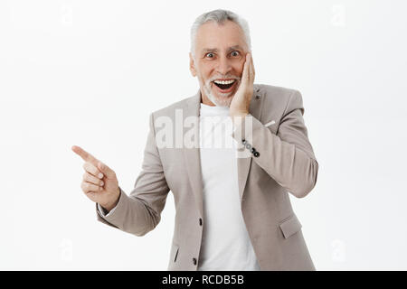 Waist-up shot of speechless delighted and surprised handsome elegant grandfather in stylish suit pressing hand to cheek from amazement and joy pointing left at curious thing smiling happily Stock Photo