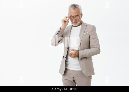 Portrait of serious good-looking wise male entrepreneur in glasses with white beard and hair touching suit and frames looking from under forehead intelligent and questioned posing over grey wall Stock Photo