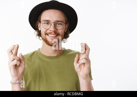 Hopeful and optimistic charming young hipster male traveler with beard in glasses and hat crossing fingers for good luck, smiling, making wish having faith dream come true, praying over gray wall Stock Photo