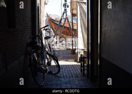 Spakenburg, Utrecht, The Netherlands: Traditional, Dutch fishing boat seen in sunlight beyond a darkened alleyway, where Dutch bikes are leaned. Stock Photo