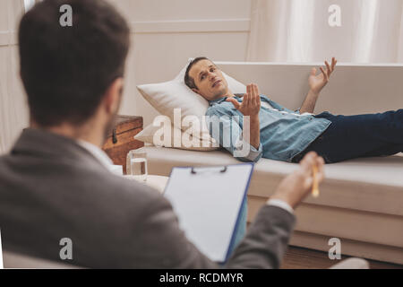 Stressed man lying on beige sofa explaining his problems to therapist Stock Photo