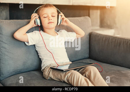 Favorite music. Adorable little boy sitting on the sofa and listening to the music in headphones from his tablet while keeping his eyes closed Stock Photo