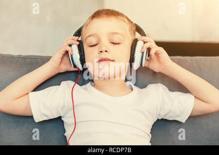 Inspiring music. Close up of a pleasant little boy sitting on the sofa and listening to the music in his headphones while keeping his eyes closed Stock Photo