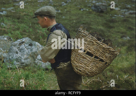1960s, historical, a man wearing a flat cap carrying a wicker basket of peat on his back, Ireland, UK. In the rural communities of Ireland, the organic material of peat was used a much needed fuel to heat their homes. This kind of wicker basket is also know as a creel and as well used for blocks of peat, was also used for carrying fish in the scottish herring industry. Stock Photo