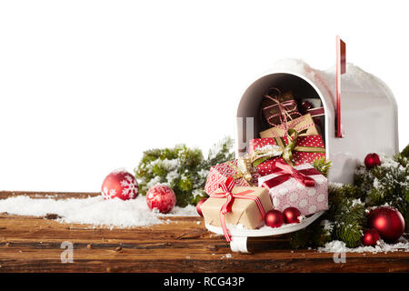 Colorful gift-wrapped Christmas presents and red baubles spilling from a letterbox onto a rustic wooden table with snow and copy space Stock Photo