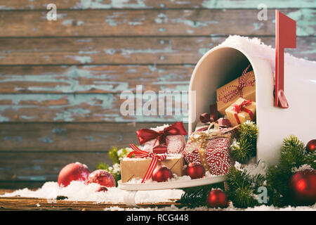 Christmas gifts and baubles spilling from a mailbox covered in white winter snow against a rustic wooden wall with copy space Stock Photo