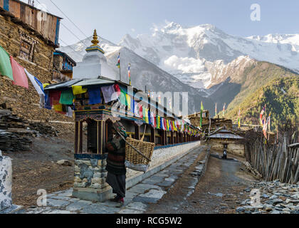Old local woman spinning prayer wheels along a Mani wall in Upper Pisang with a snow avalanche on Annapurna II mountain in the background, Nepal Stock Photo