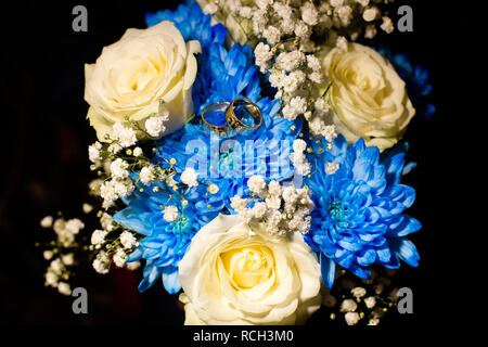 A pair of wedding gold rings on a bouquet of white and blue flowers on a black background. Shot close up. Stock Photo