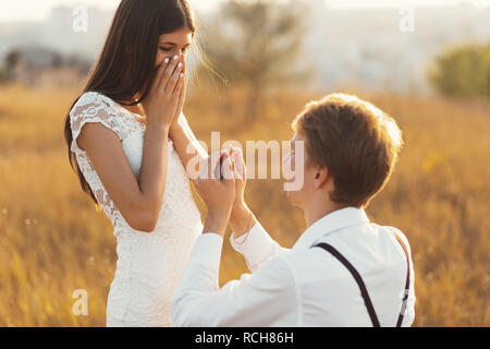 The Unique moment in their life, young man standing at his knee proposing her. She is happy, can't believe her eyes, covering face with hands, on a go Stock Photo