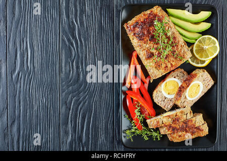 meatloaf with hard boiled egg filling served with red pepper and avocado slices on a black rectangular platter on a wooden table, view from above, fla Stock Photo