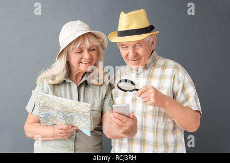 Senior couple tourists wearing beach hats studio standing isolated on gray wall holding map looking at smartphone through magnifying glass browsing in Stock Photo